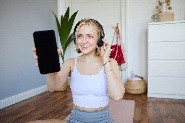 Ritratto di una donna in forma e in salute che si allena a casa indossando le cuffie che mostra lo schermo dello smartphone vuoto