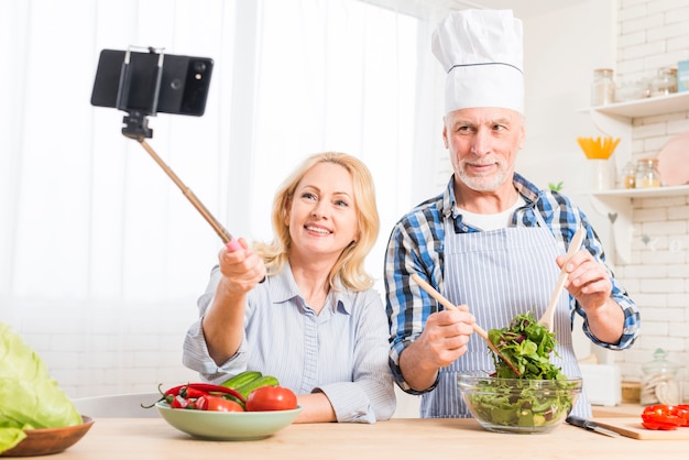 Ritratto di una donna senior che prende selfie sul telefono cellulare con il suo marito che prepara l&#39;insalata nella cucina