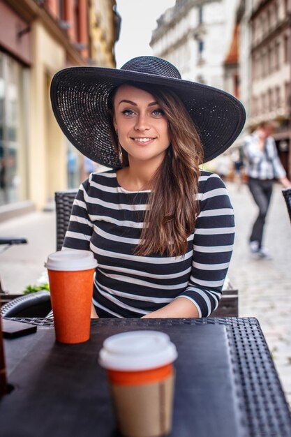 Ritratto di una donna sorridente che indossa un vestito e un cappello alla moda seduto in un caffè di strada estivo.