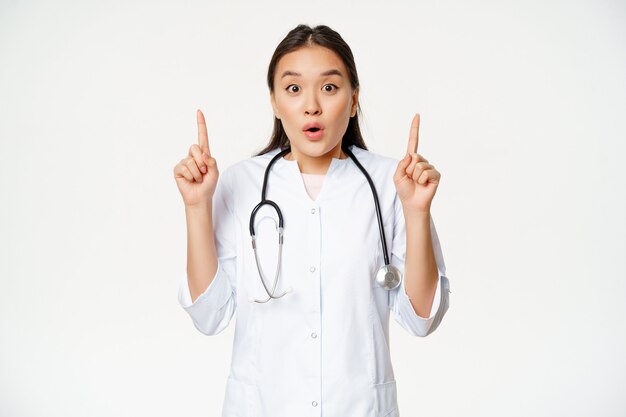 Ritratto di una dottoressa asiatica sorpresa, medico che punta le dita in alto, mostrando notizie straordinarie, grande promo, sfondo bianco.
