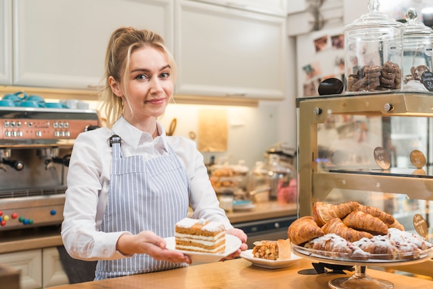 Ritratto di una fetta di torta bionda sorridente della tenuta della giovane donna sul piatto nella caffetteria