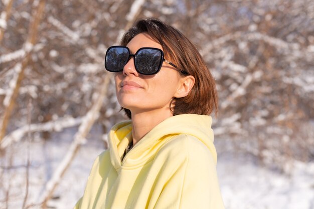 Ritratto di una giovane bella donna in una foresta di inverno paesaggio innevato in una giornata di sole, vestita con un grande pullover giallo, con occhiali da sole, godersi il sole e la neve