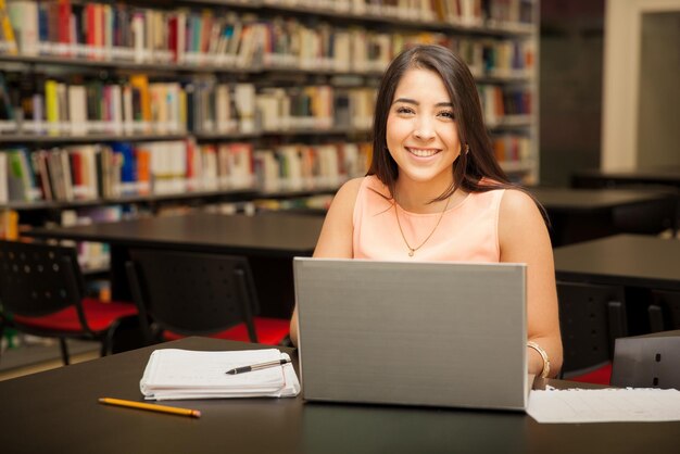 Ritratto di una giovane bruna che utilizza un computer portatile per il lavoro scolastico in biblioteca e sorridente