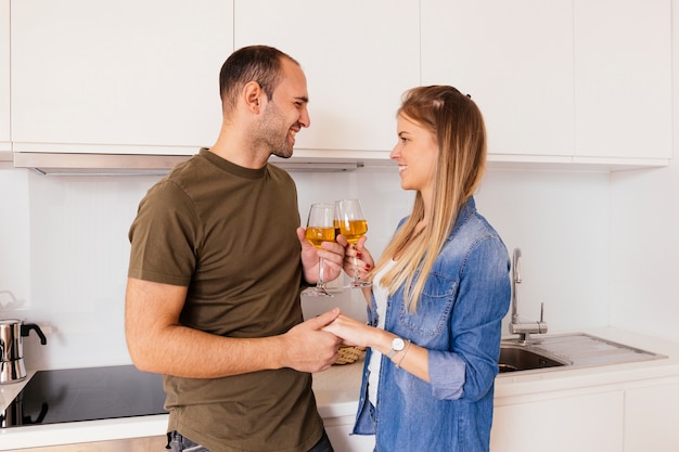 Ritratto di una giovane coppia sorridente che tiene la mano di ciascuno che tosta i bicchieri di vino nella cucina