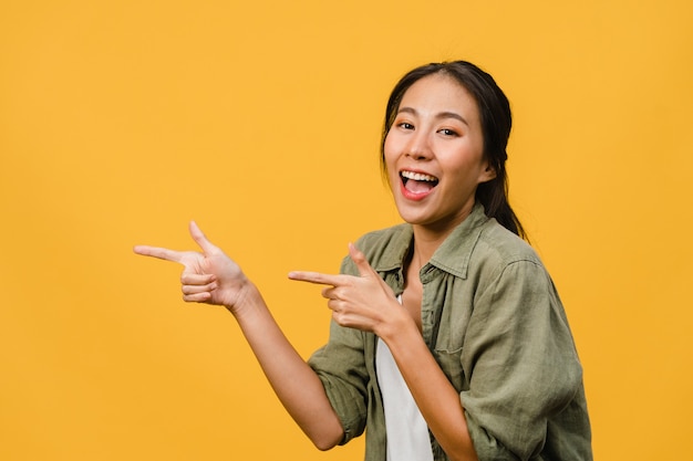 Ritratto di una giovane donna asiatica che sorride con un'espressione allegra, mostra qualcosa di straordinario in uno spazio vuoto in un panno casual isolato su un muro giallo. Concetto di espressione facciale.
