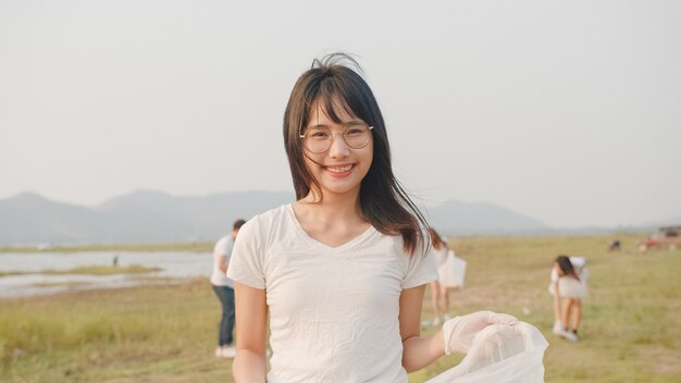 Ritratto di una giovane donna asiatica volontaria aiuta a mantenere pulita la natura, guardando davanti e sorridendo con i sacchi della spazzatura bianchi sulla spiaggia
