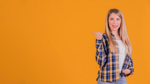 Ritratto di una giovane donna che guarda e che punta al lato con il pollice contro uno sfondo arancione