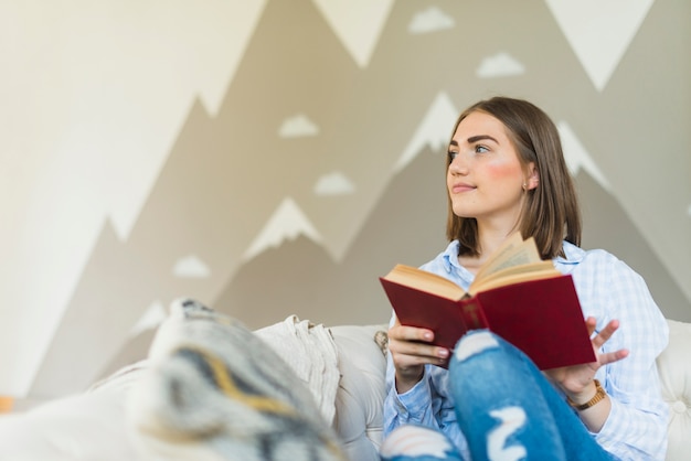 Ritratto di una giovane donna che si siede sul divano con la holding del libro