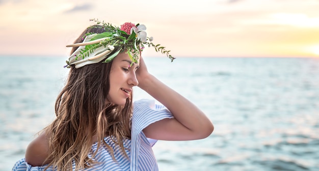 Ritratto di una giovane donna con una composizione di fiori sulla sua testa sullo sfondo dell'oceano al tramonto.