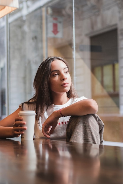 Ritratto di una giovane donna contemplata che tiene sguardo disponibile della tazza di caffè eliminabile