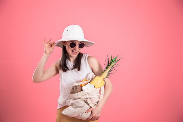 Ritratto di una giovane donna elegante vestita in abiti estivi con un cappello e occhiali da sole, in possesso di un sacchetto di frutta ECO, in rosa isolato.