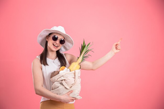 Ritratto di una giovane donna elegante vestita in abiti estivi con un cappello e occhiali da sole, in possesso di un sacchetto di frutta ECO, in rosa isolato.