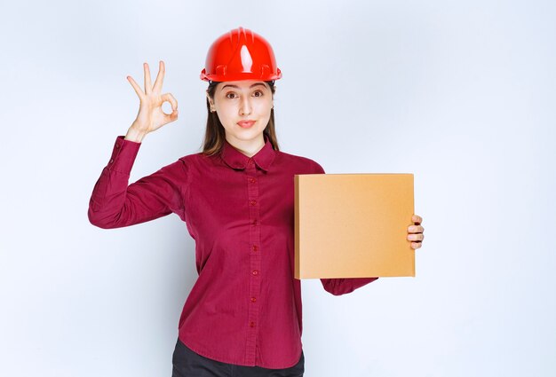 Ritratto di una giovane donna in casco con piccola scatola di carta che mostra gesto ok.