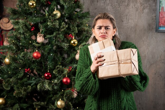 Ritratto di una giovane donna in maglione verde che tiene pila di scatole regalo e guardando la fotocamera