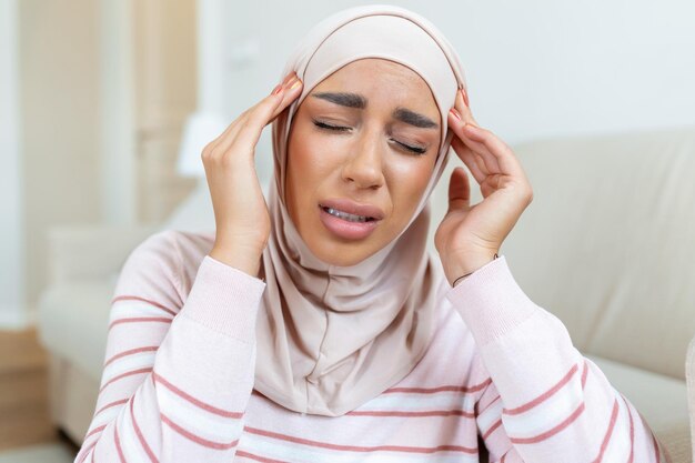 Ritratto di una giovane donna musulmana araba seduta sul divano di casa con mal di testa e dolore Donna con hijab che soffre di mal di testa cronico quotidiano Donna triste che tiene la testa a causa del dolore al seno