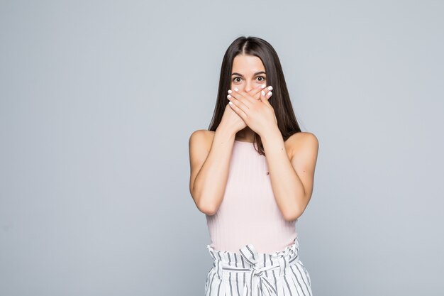 Ritratto di una giovane donna sorpresa che copre la bocca con le mani isolate sul muro bianco