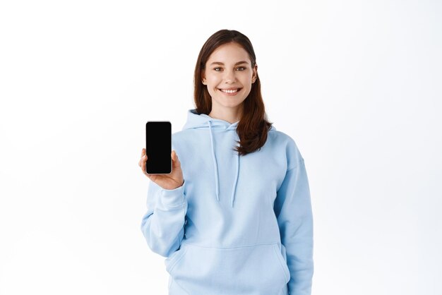 Ritratto di una giovane donna sorridente che mostra il telefono cellulare in bianco dello schermo isolato sopra priorità bassa bianca