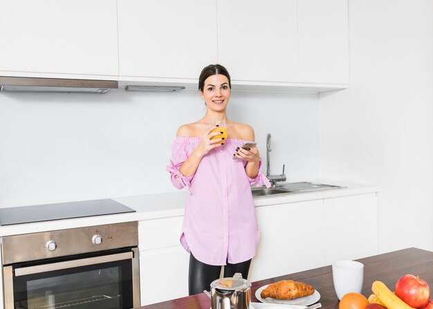 Ritratto di una giovane donna sorridente che tiene vetro di succo e telefono cellulare in cucina