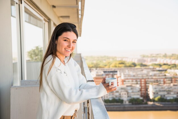 Ritratto di una giovane donna sorridente in piedi in balcone tenendo tazza di caffè bianco