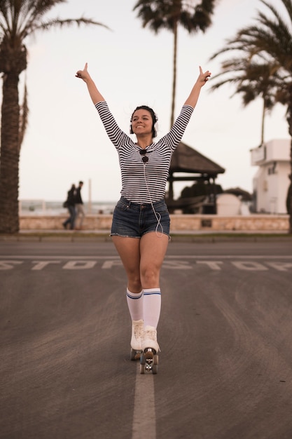 Ritratto di una giovane donna sorridente in piedi sulla strada alzando le sue mani ascolto musica in cuffia