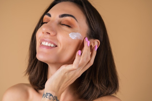 Ritratto di una giovane donna su una parete beige con un trucco naturale e caldo e una pelle liscia e pulita, applica la crema sul viso