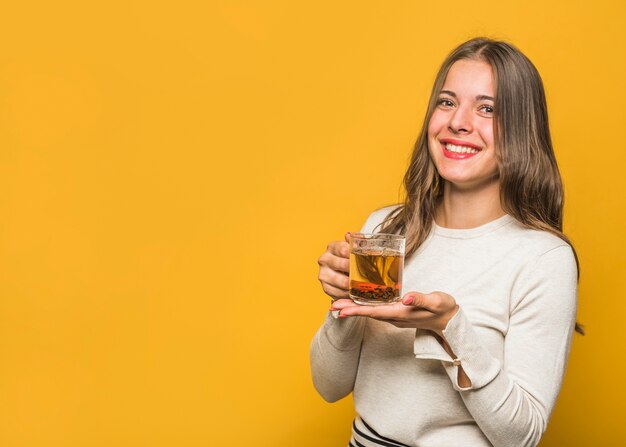 Ritratto di una giovane e bella donna che mostra tazza di vetro tisana contro sfondo giallo