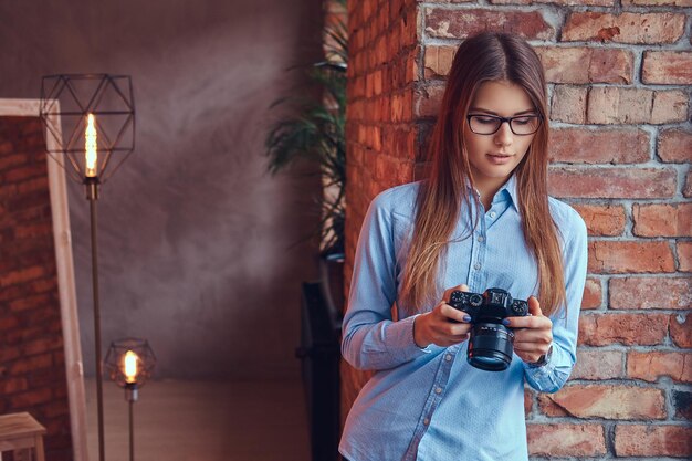 Ritratto di una giovane fotografa in occhiali e camicia blu appoggiata a un muro di mattoni in una stanza con design loft.