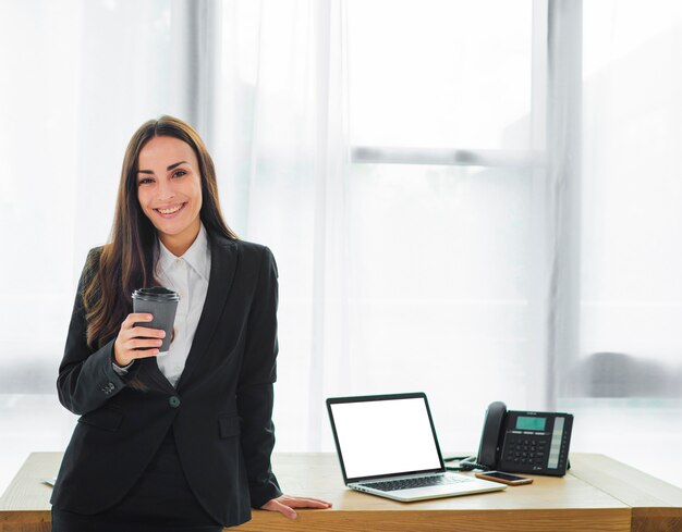 Ritratto di una giovane imprenditrice sorridente in piedi davanti alla scrivania tenendo la tazza di caffè usa e getta