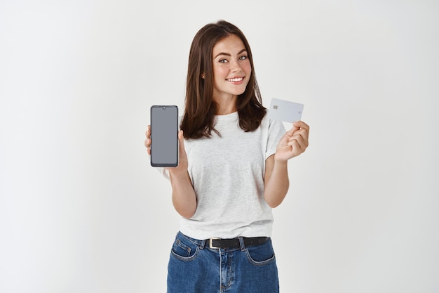 Ritratto di una giovane ragazza felice con lunghi capelli castani in piedi sul muro bianco, che mostra una carta di credito in plastica e uno schermo vuoto dello smartphone.