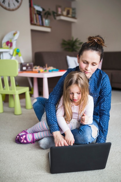 Ritratto di una madre con sua figlia seduta sul tappeto con laptop