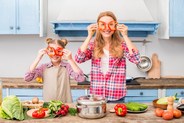 Ritratto di una madre e una figlia sorridenti che tengono la fetta del peperone davanti agli occhi che esaminano macchina fotografica