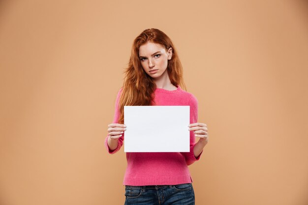 Ritratto di una ragazza abbastanza arrabbiata di redhead che mostra cartello bianco in bianco