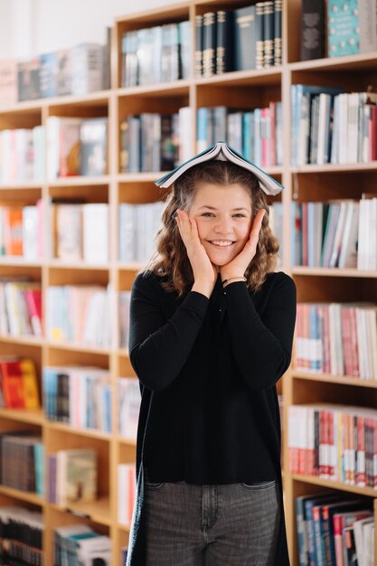 Ritratto di una ragazza adolescente caucasica sorridente divertente che mette e tiene in mano un libro sulla testa e guarda la fotocamera, sfondo di scaffali.