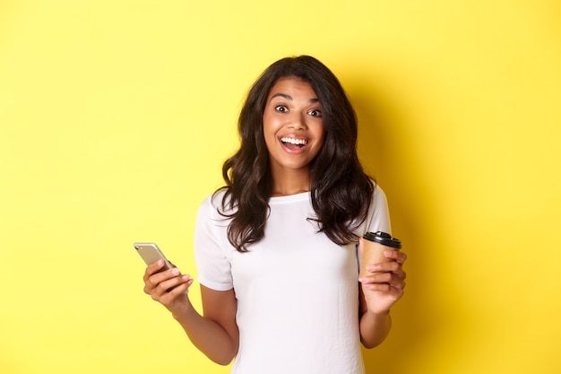 Ritratto di una ragazza afroamericana attraente che sorride, tiene in mano una tazza di caffè e uno smartphone, in piedi su sfondo giallo