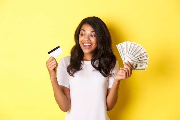 Ritratto di una ragazza afroamericana attraente che tiene in mano soldi e guarda la carta di credito in piedi
