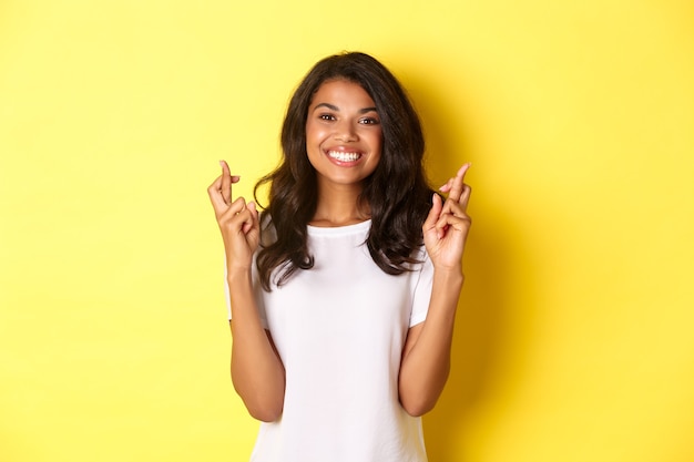 Ritratto di una ragazza afroamericana ottimista e sorridente, che incrocia le dita per buona fortuna e fa un desiderio, in piedi su sfondo giallo.