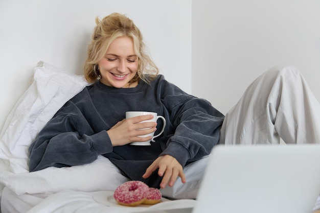 Ritratto di una ragazza bionda felice modello femminile sdraiata a letto in abiti accoglienti mangiando e guardando un film