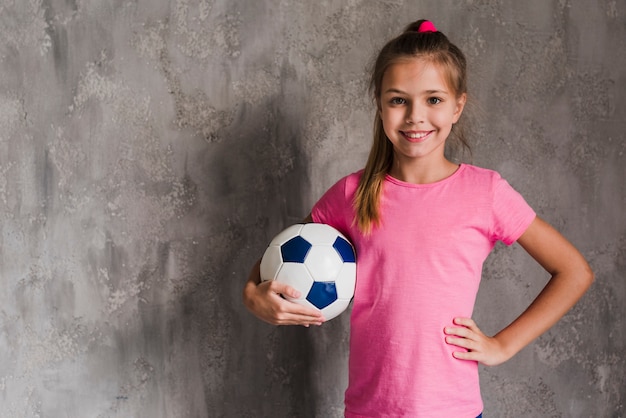 Ritratto di una ragazza bionda sorridente con la mano sul pallone da calcio della tenuta dell&#39;anca contro la parete grigia