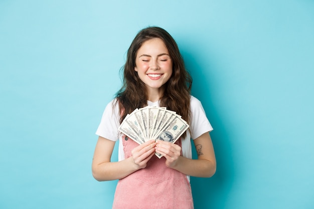 Ritratto di una ragazza carina che sorride con soddisfazione, tiene in mano soldi e sembra soddisfatta, vincendo un premio in banconote da un dollaro, in piedi su sfondo blu.