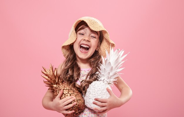 Ritratto di una ragazza carina in un cappello estivo, tenendo in mano due ananas dipinti in oro e bianco, il concetto di estate e creatività