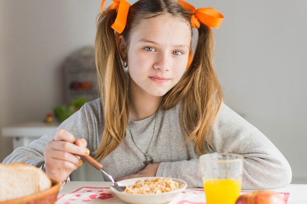Ritratto di una ragazza che mangia i cereali sani con bicchiere di succo sul tavolo