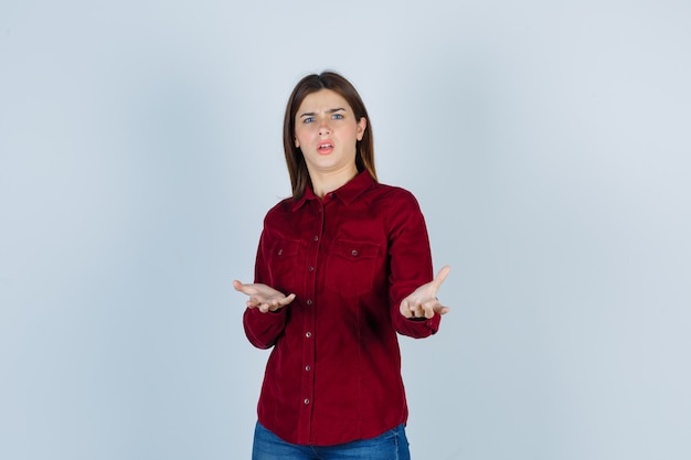 Ritratto di una ragazza che mostra un gesto di domanda in camicia bordeaux e sembra perplessa