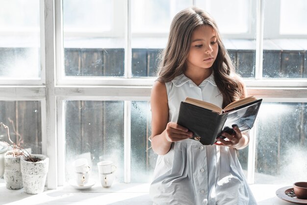 Ritratto di una ragazza che si siede davanti al libro di lettura della finestra