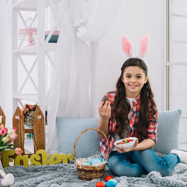 Ritratto di una ragazza che si siede sul letto che tiene l&#39;uovo di cioccolato di Pasqua e ciotola di caramelle che guarda alla macchina fotografica