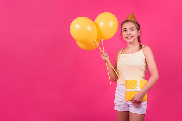 Ritratto di una ragazza felice che tiene scatola regalo giallo e palloncini in mano su sfondo rosa