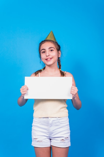 Ritratto di una ragazza in cappello di compleanno che mostra carta bianca in piedi su sfondo blu