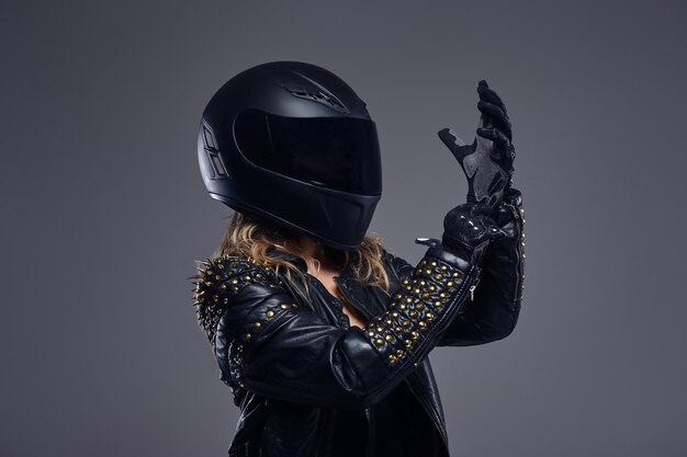 Ritratto di una ragazza motociclista che indossa un costume da corsa in pelle e un casco protettivo che indossa i guanti in uno studio su sfondo grigio.
