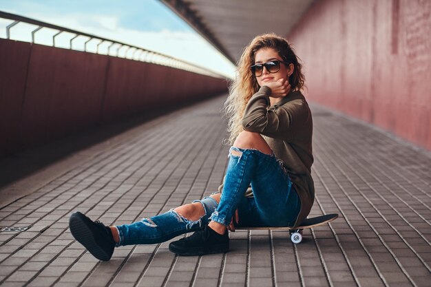 Ritratto di una ragazza pensierosa in occhiali da sole vestita con una felpa con cappuccio e jeans strappati seduta su uno skateboard su un marciapiede del ponte.