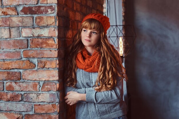 Ritratto di una ragazza rossa solitaria che indossa un maglione caldo e un cappello con le braccia incrociate appoggiata a un muro di mattoni.