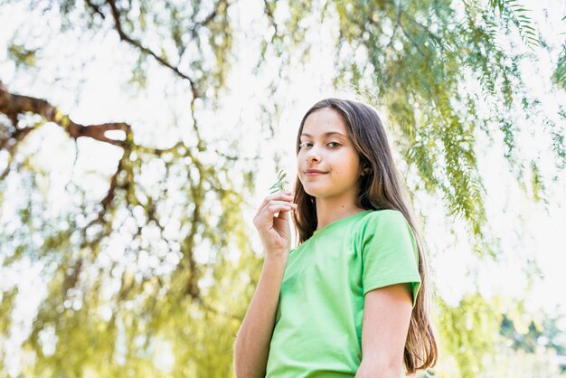 Ritratto di una ragazza sorridente che giudica disponibile della felce che esamina macchina fotografica che sta sotto l&#39;albero